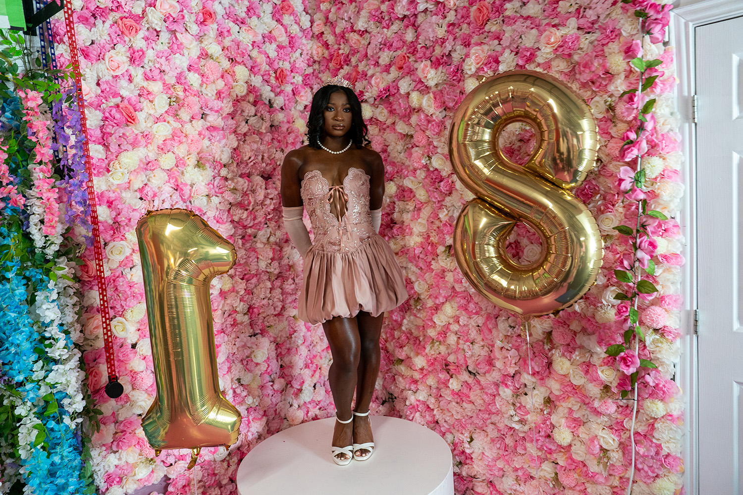 A young woman in a pink dress stands on a white platform between gold balloons shaped as the number 18, with a pink floral wall as the backdrop.