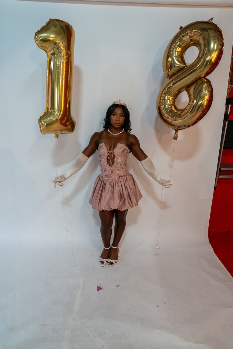 A person in a pink dress and tiara holds gold balloons shaped as the number 18, standing against a white backdrop.