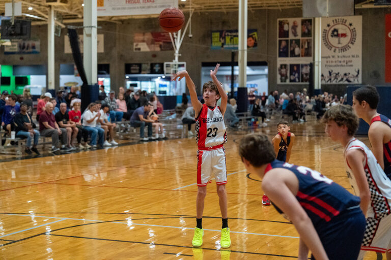 A youth basketball player in a white and red uniform takes a free throw while other players and spectators watch in a gymnasium.