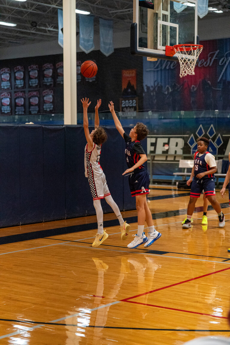 Two youth basketball players jump near the hoop as one attempts a shot while the other defends; other players and banners are visible in the background.