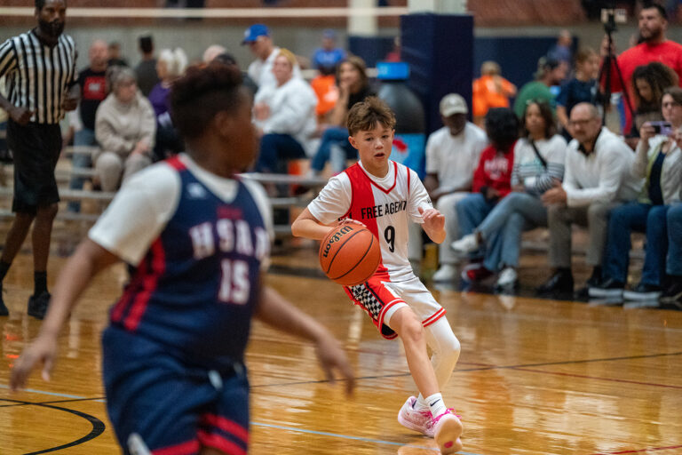 A youth basketball player in a white "Free Agents" jersey dribbles the ball while being guarded by an opposing player in a blue "Huskies" jersey, with spectators watching in the background.