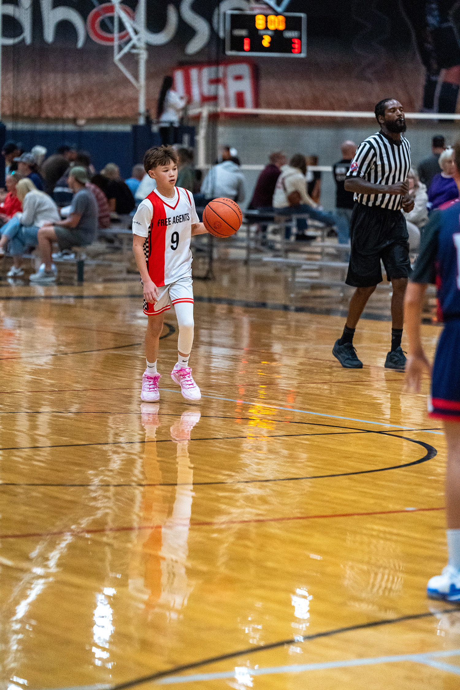 A youth basketball player dribbles the ball on an indoor court during a game, with a referee and spectators in the background.