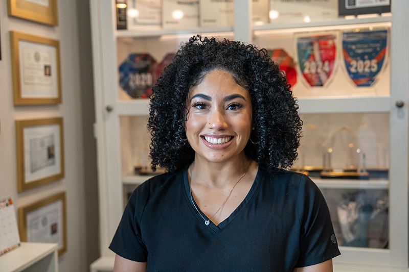 A woman with curly hair wearing a black scrub top smiles at the camera, standing in front of a display case with awards and certificates.