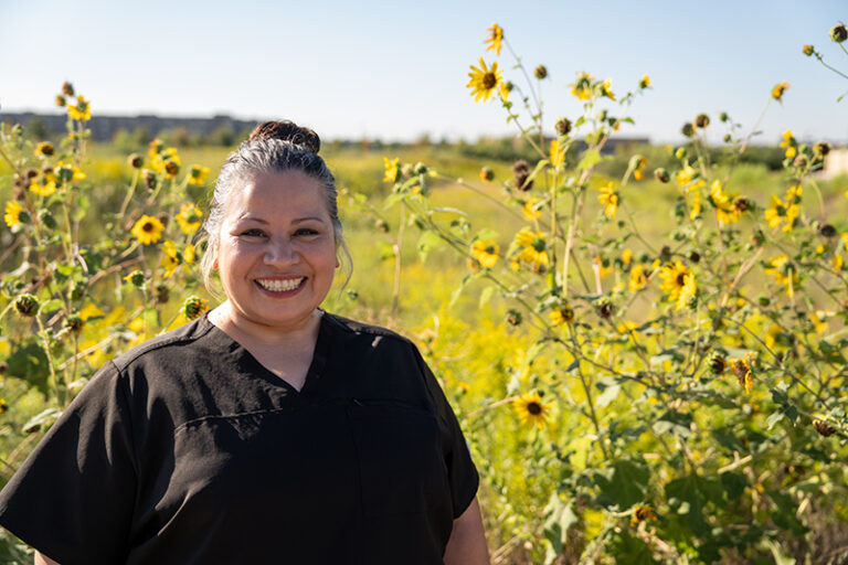 Woman in black scrubs smiles while standing outdoors in a field with blooming yellow sunflowers under a clear sky.