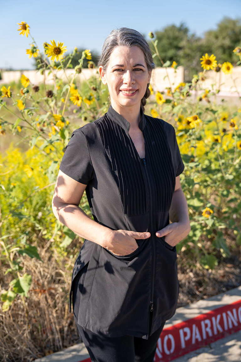 A woman with gray hair pulled back, wearing a black zip-up top, stands outdoors in front of sunflowers and a "No Parking" curb sign, smiling at the camera.