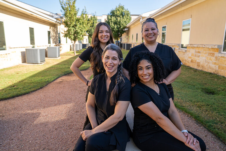 Four women in black scrubs smile outdoors near a building on a sunny day, with two sitting on a low wall and two standing behind them.