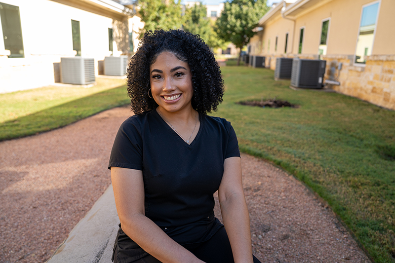 A woman with curly hair wearing a black shirt sits outside on a stone border, smiling at the camera with buildings and grass in the background.