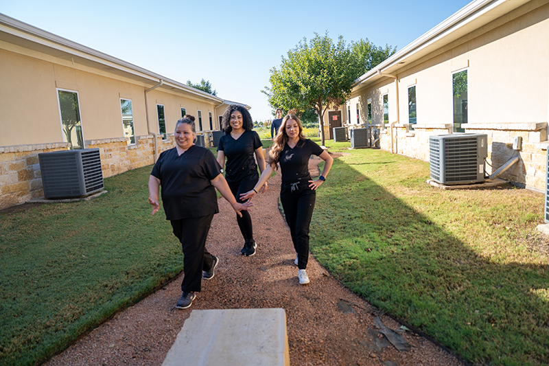Three women in black scrubs walk on a gravel path between two beige buildings with air conditioning units on a sunny day.