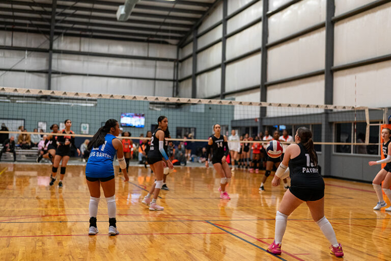 A women's indoor volleyball game in progress, with players in action near the net and spectators seated in the background.