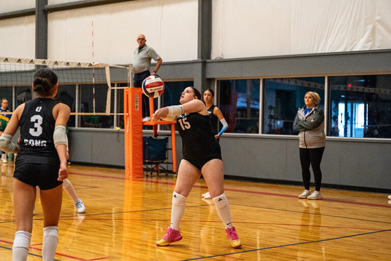 A volleyball player in a black jersey prepares to receive the ball during an indoor match, while teammates and coaches watch from the sidelines.