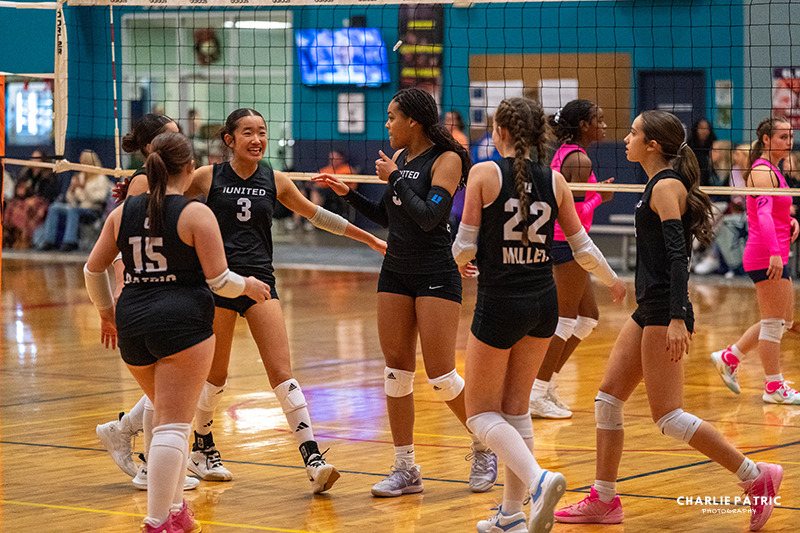 A girls' volleyball team in black uniforms gathers and smiles on an indoor court during a game, with spectators visible in the background—capturing the spirit of youth sports photography in Frisco.