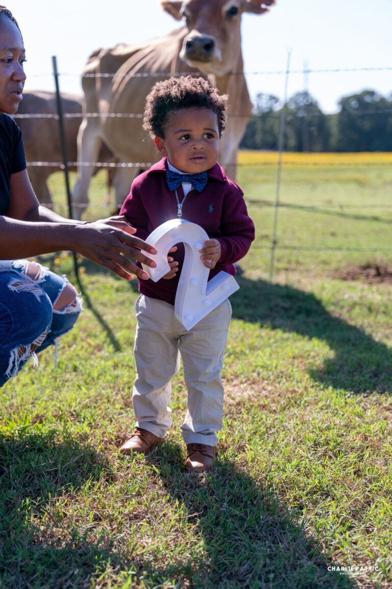 A young boy stands on grass holding a large number two, with an adult nearby and a cow behind a fence—capturing moments like these is why knowing how to choose an event photographer is so important.