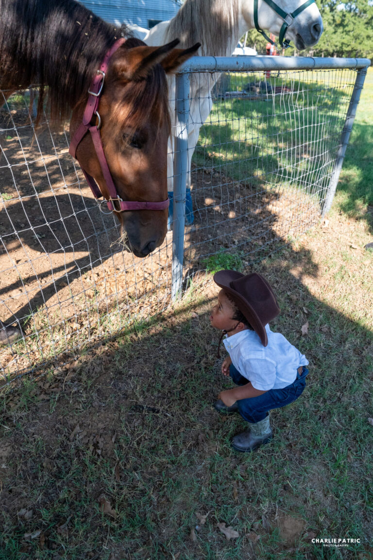 A young child in cowboy attire crouches near a fence, looking at a brown horse with a pink halter. Capturing candid moments like these is one of the best event photography tips for success. Another white horse stands in the background.