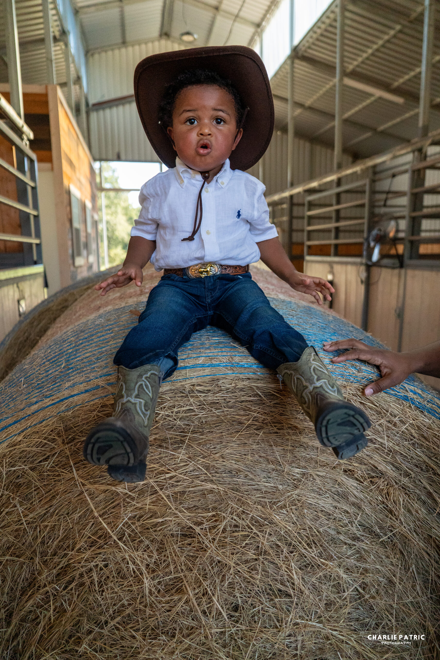A young child in a cowboy hat, boots, and a white shirt sits atop a large hay bale inside a barn—perfect inspiration for Frisco event photography venues—with an adult’s hands nearby for support.