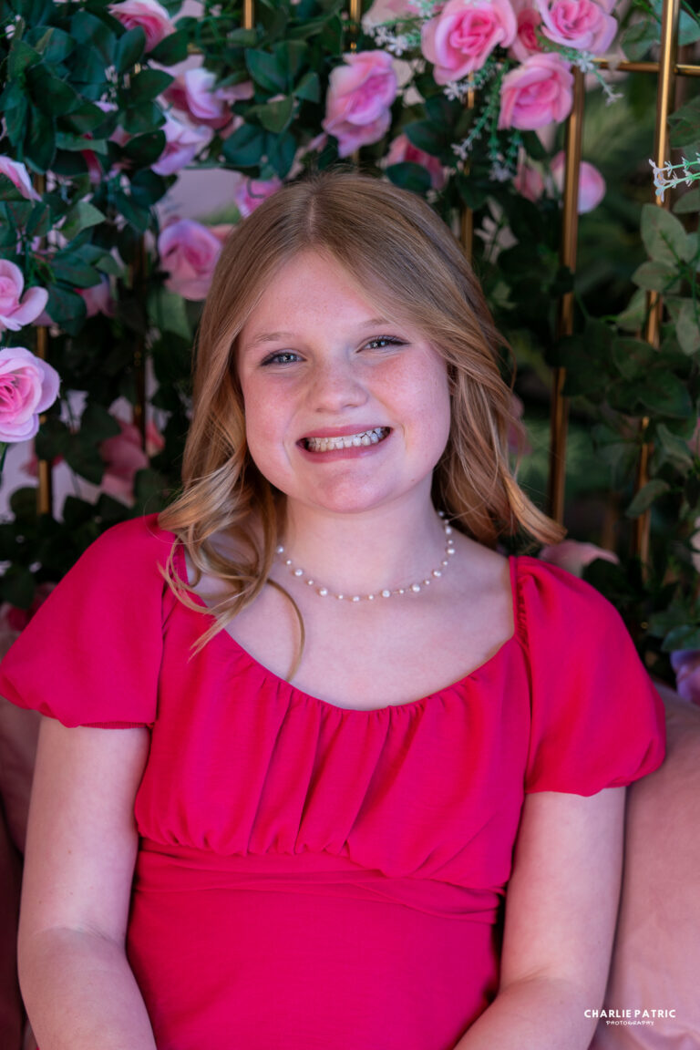 A young girl with blonde hair, wearing a bright pink dress and a pearl necklace, smiles while sitting in front of green leaves and pink roses, beautifully highlighting the impact of lighting in event photography.