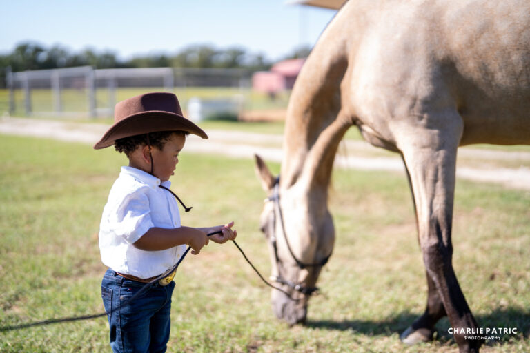 A young child wearing a cowboy hat stands outdoors holding a rope near a light brown horse grazing on grass, capturing a scene reminiscent of how to choose an event photographer who can preserve such special moments.