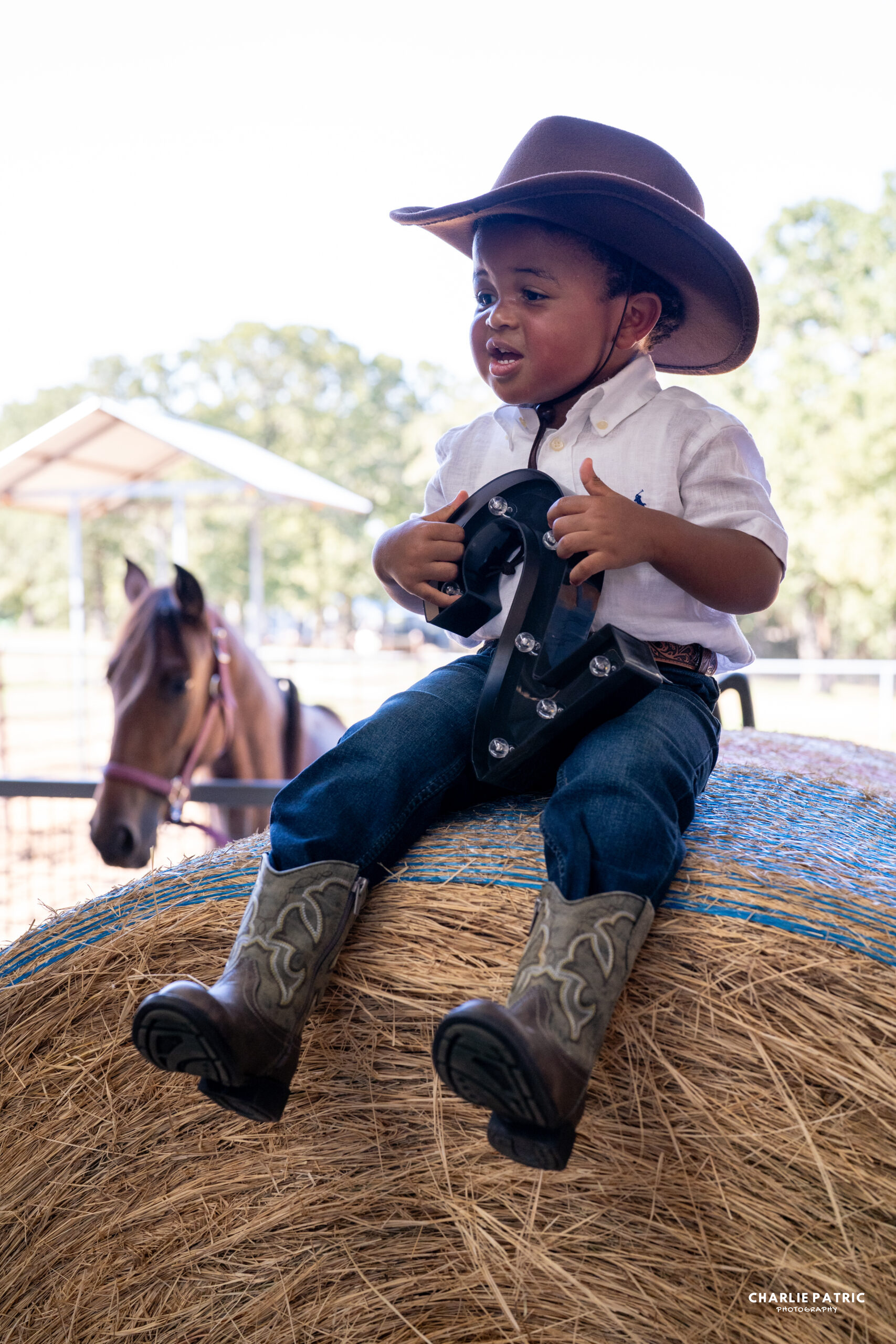 Young boy in a cowboy hat and boots sits on a hay bale, holding a toy horse saddle; a horse stands in the background, perfectly capturing emotions in event photography.