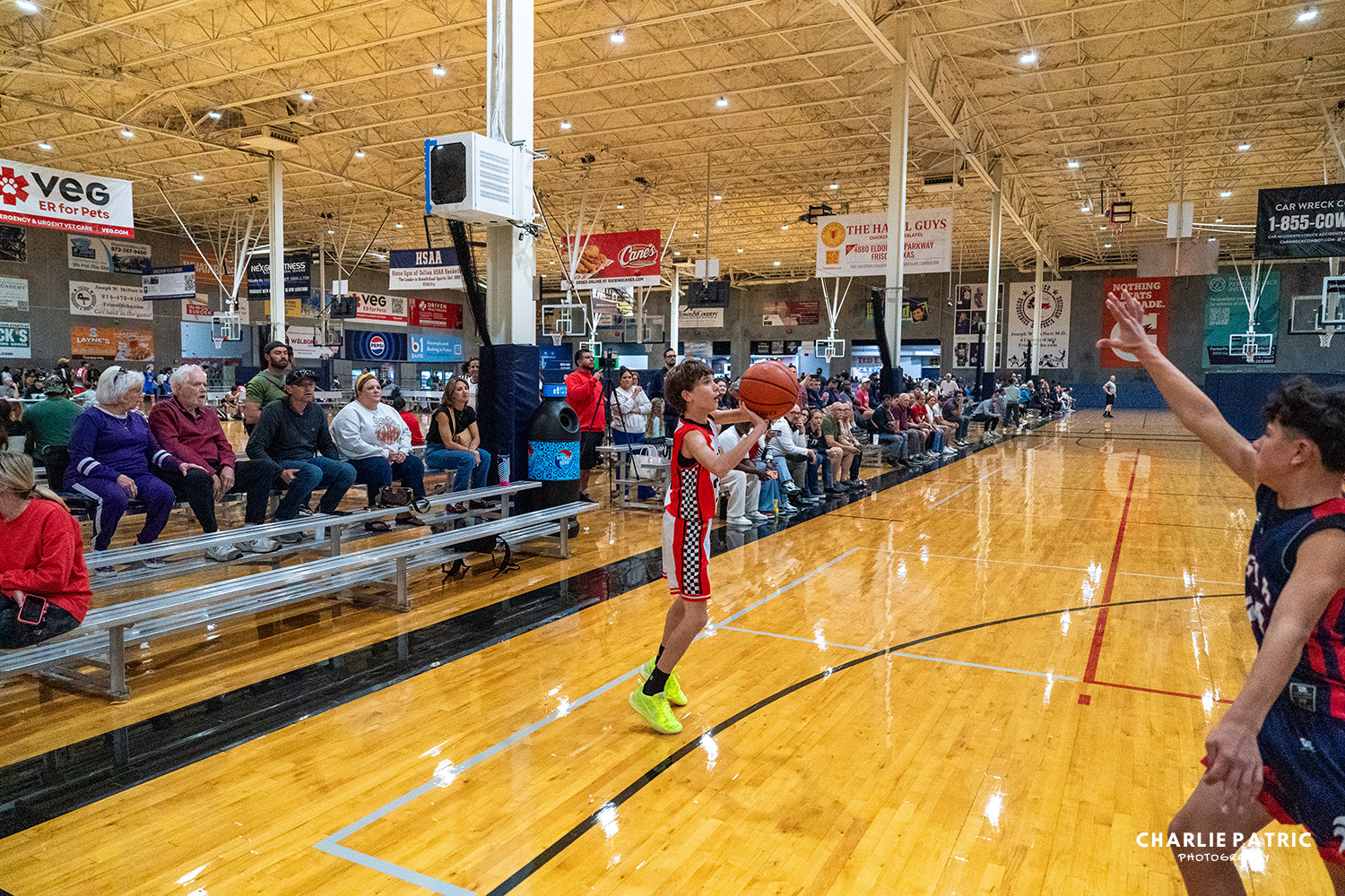 A youth basketball player in a red and black uniform prepares to shoot while a defender raises their arm; spectators watch from the bleachers in a gymnasium, capturing an exciting moment of youth sports photography in Frisco.
