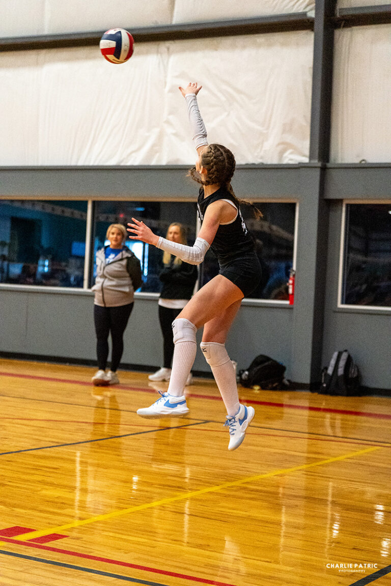 Capturing Youth Sports Action, a volleyball player leaps mid-air to serve the ball in an indoor gym, while three people watch from the sidelines.