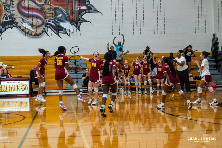 A girls' volleyball team in maroon uniforms celebrates on the court, captured vividly through the best lenses, while teammates and supporters cheer from the bench and bleachers.