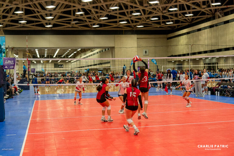 Indoor volleyball match in progress; one player in a red uniform jumps to hit the ball while teammates and opposing team members prepare to react. Spectators watch from the background, making it a great setting to test the best lenses for sports photography.