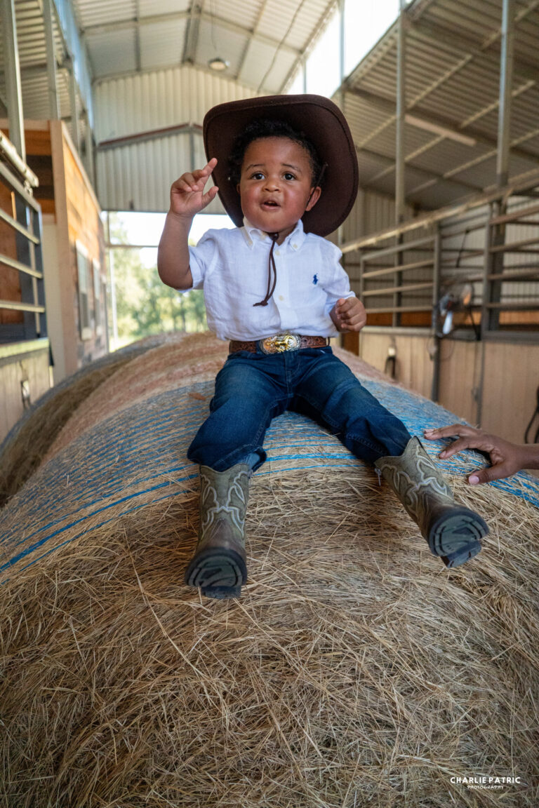 A young child wearing a cowboy hat, white shirt, jeans, and boots sits on a large hay bale inside a barn, holding up two fingers—showcasing how the impact of venue on event photography creates authentic moments.