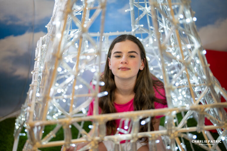 A young woman with long brown hair and a pink shirt sits inside a structure decorated with white string lights, against a sky-themed backdrop—perfect for capturing high-resolution images.
