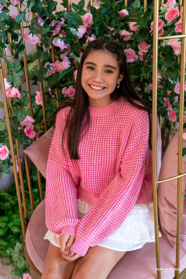 A young woman with long dark hair, wearing a pink sweater and white skirt, sits on a pink chair surrounded by green leaves and pink roses, smiling at the camera, showcasing the impact of venue on event photography.