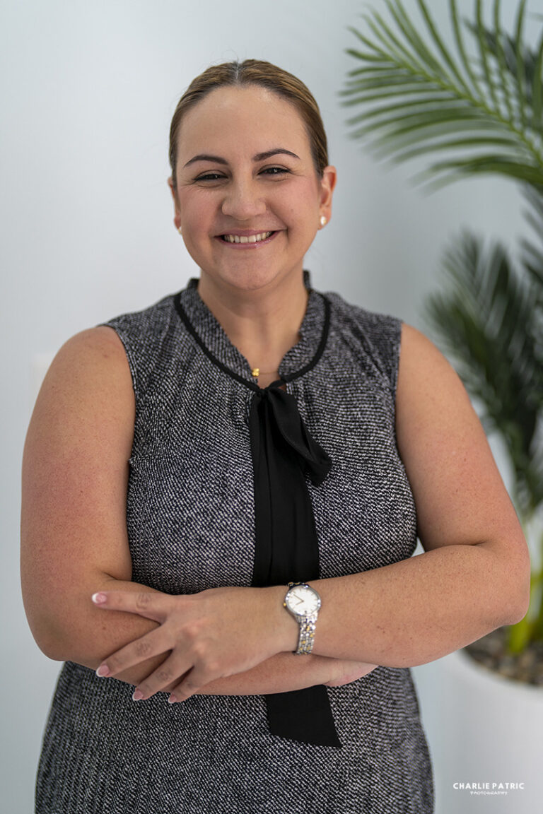 A woman with light brown hair in a ponytail, wearing a sleeveless grey dress with a black bow, stands smiling with arms crossed in front of a leafy plant, highlighting the impact of background on the photo's atmosphere.