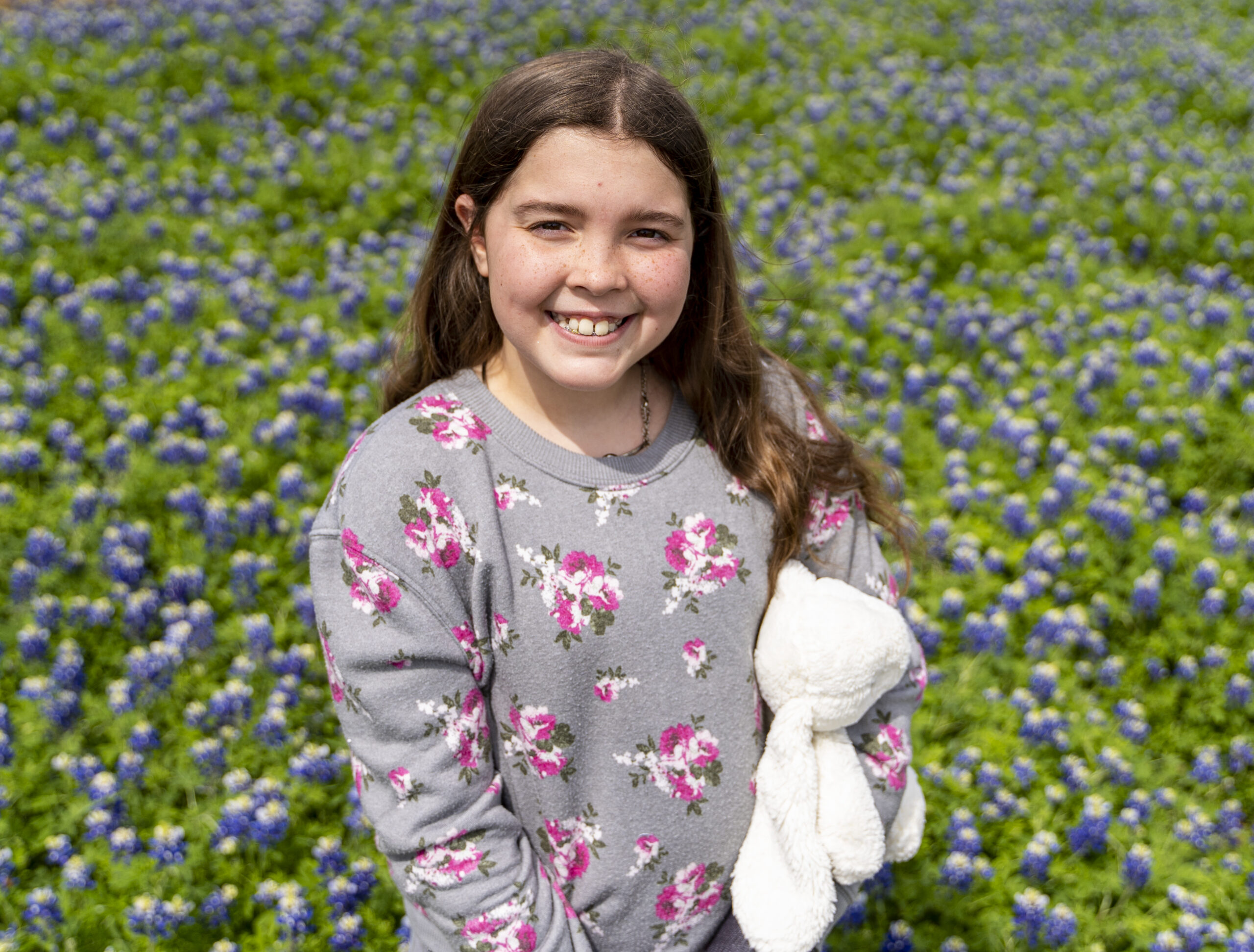 Smiling girl with long brown hair holding a white stuffed animal, standing in a field of blue and green wildflowers during a bluebonnet photoshoot in Frisco, TX.