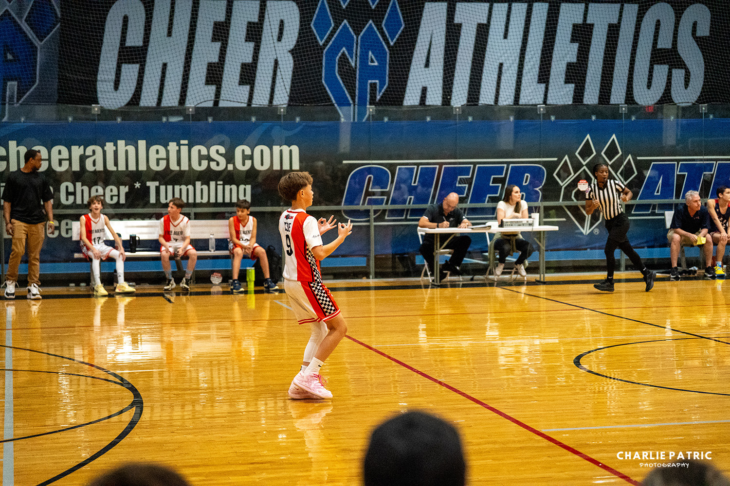 A youth basketball player in a red and white uniform holds the ball on a court. Captured using the best camera settings for sports events, teammates and coaches are seen seated on the bench in the background.