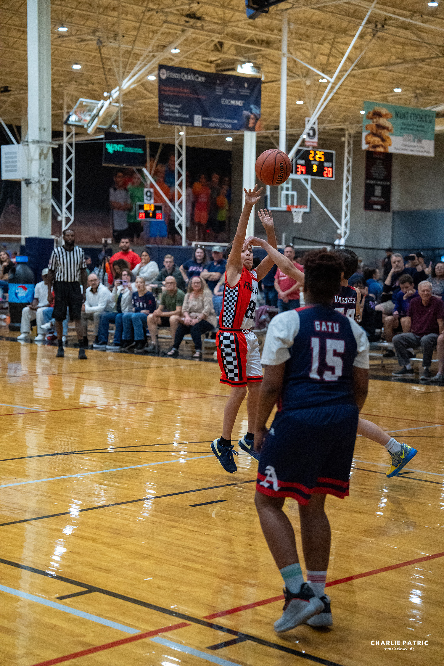 A youth basketball player in a red jersey leaps to shoot as a defender in blue challenges on an indoor court—spectators watch closely from the bleachers, capturing action like this with the best camera settings for sports events.