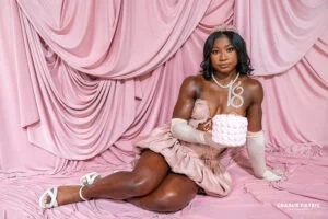 A woman in a pink dress, tiara, and gloves poses on a pink draped backdrop, holding a small pink cake with a “16” topper—perfect inspiration for personalized graduation photography.