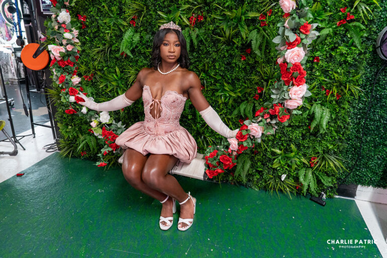 A woman in a pink dress and tiara sits on a swing adorned with red and white roses, set against a green leafy wall—one of the most enchanting Frisco TX graduation portrait locations.