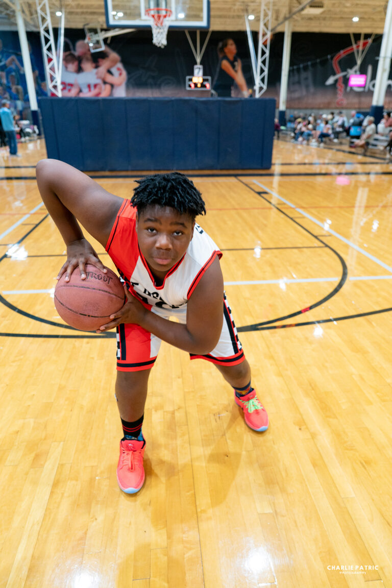 A basketball player in uniform poses in a defensive stance while holding a basketball on an indoor court, showcasing the power of editing sports photos for impact.