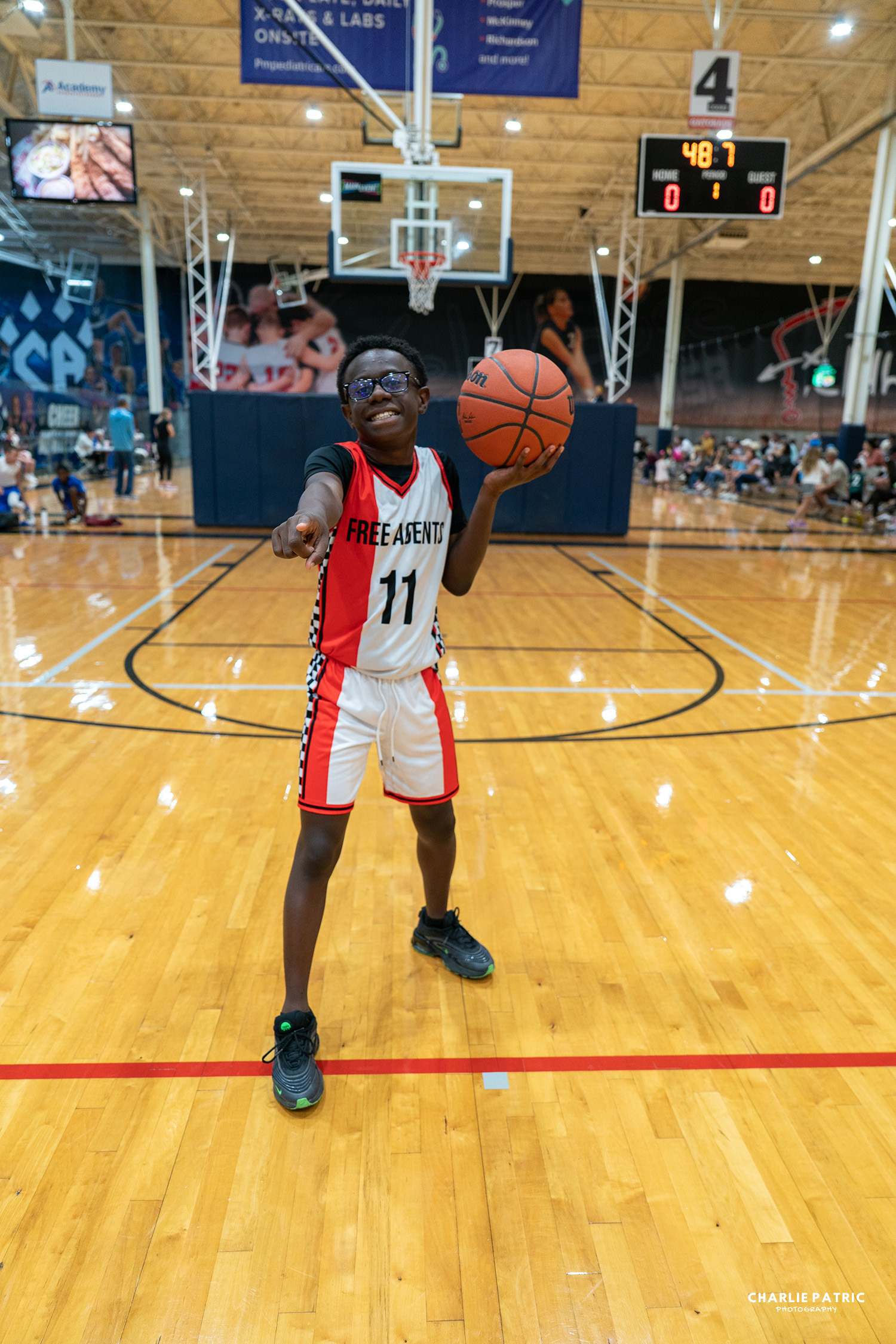 Capturing youth sports action, a young basketball player wearing a "Free Agents" jersey holds a basketball and points forward on an indoor court.