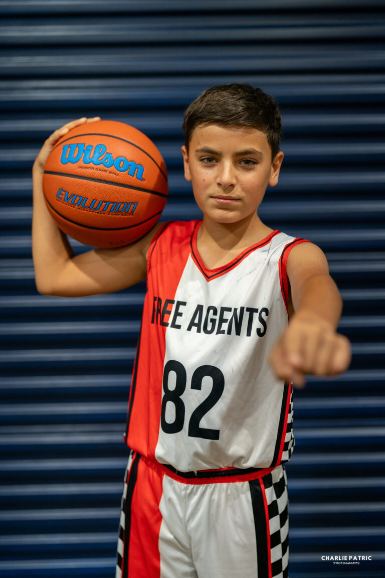 A boy in a "Free Agents 82" basketball uniform holds a Wilson basketball under one arm and points forward, standing in front of a blue corrugated metal background—perfectly captured with the best lenses for sports photography.