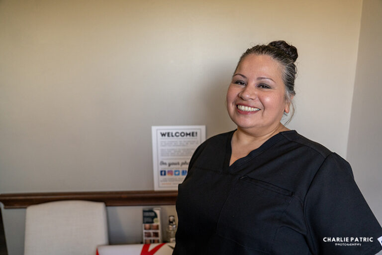 A woman in black scrubs stands indoors, smiling at the camera, as if ready for professional headshots. A welcome sign and chair are visible in the background.