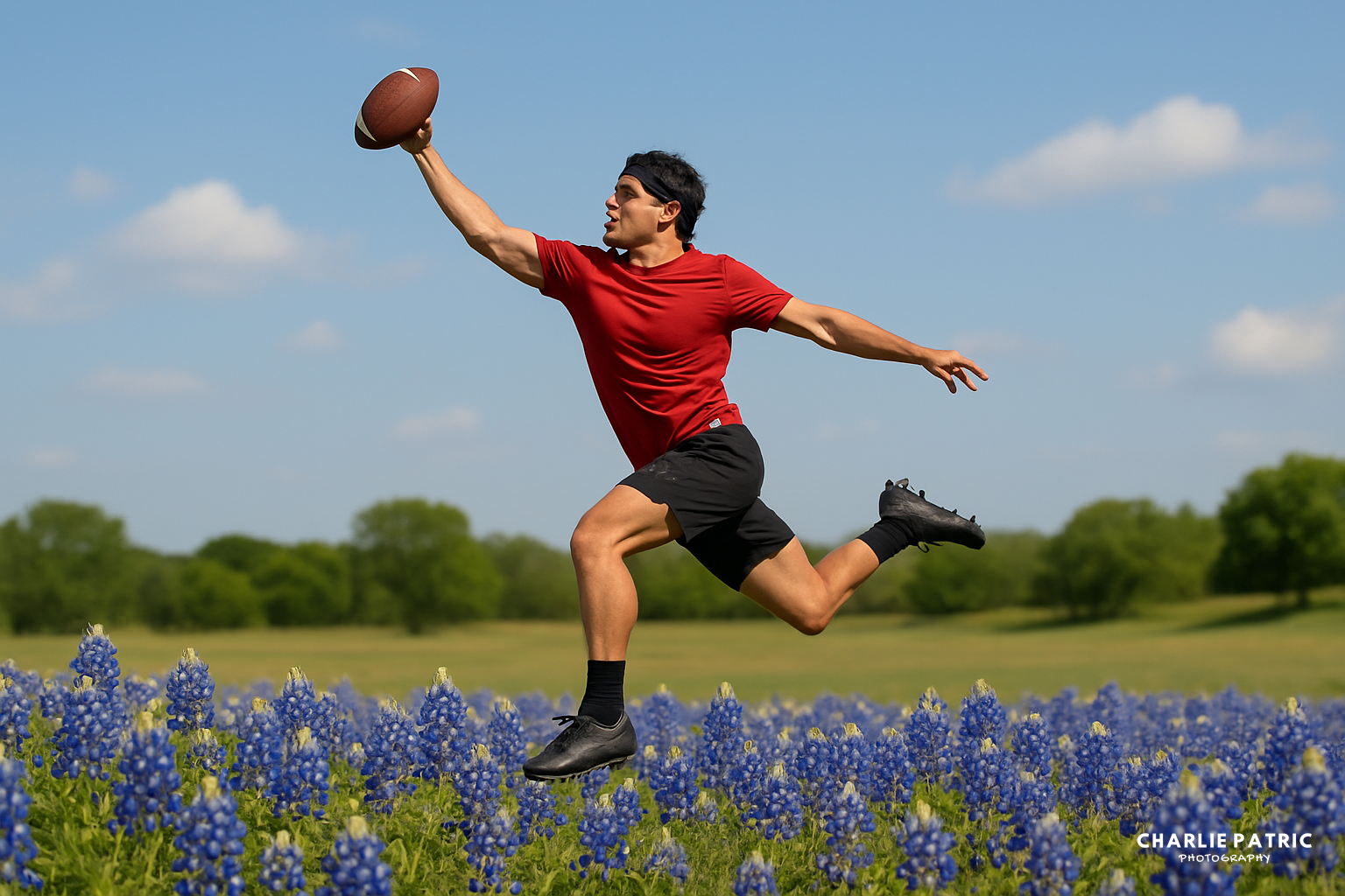 A man in a red shirt and black shorts leaps to catch a football amid vibrant bluebonnet flowers under a clear sky.