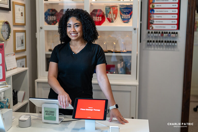 A woman stands behind a counter in a professional office, smiling beside a point-of-sale system and a tablet displaying "Folio Massage Therapy," creating an ideal setting for professional headshots or updating your LinkedIn profile.