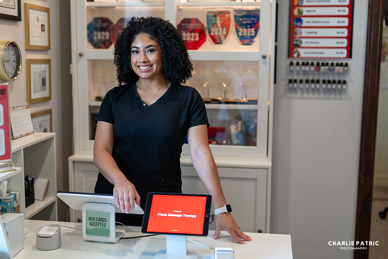 A woman stands behind a counter in a professional office, smiling beside a point-of-sale system and a tablet displaying "Folio Massage Therapy," creating an ideal setting for professional headshots or updating your LinkedIn profile.