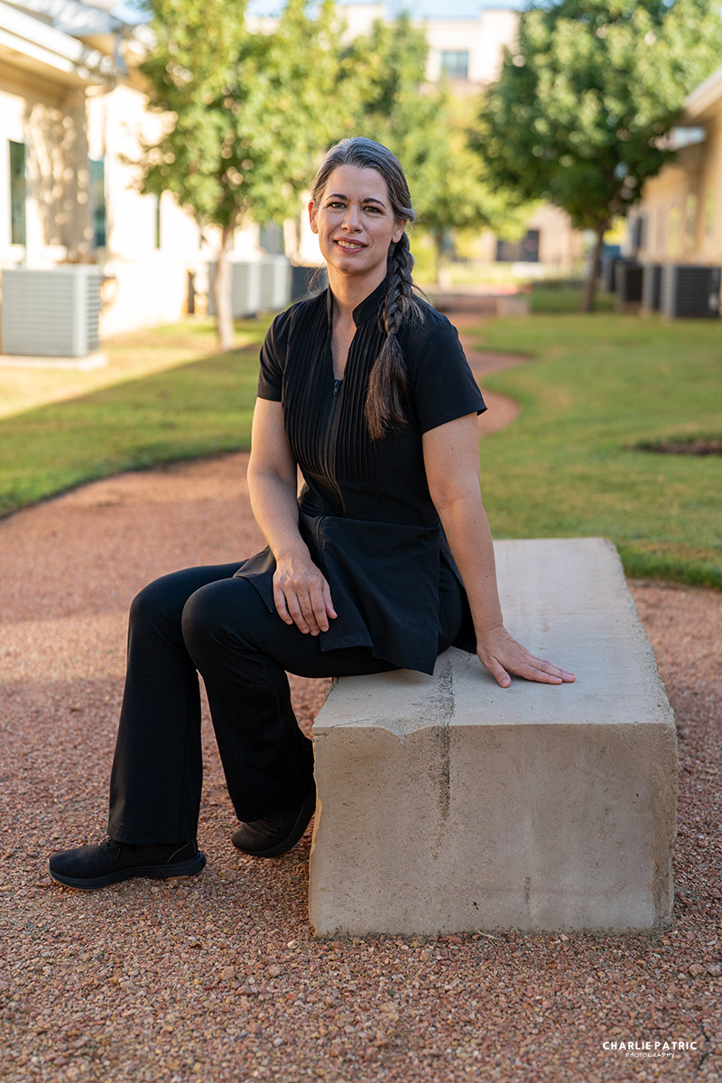 A woman with a braid, dressed in black, sits on a concrete bench along a gravel path in a landscaped outdoor area—capturing the professional photo impact often seen in corporate headshots Frisco. Trees and buildings create an elegant backdrop.