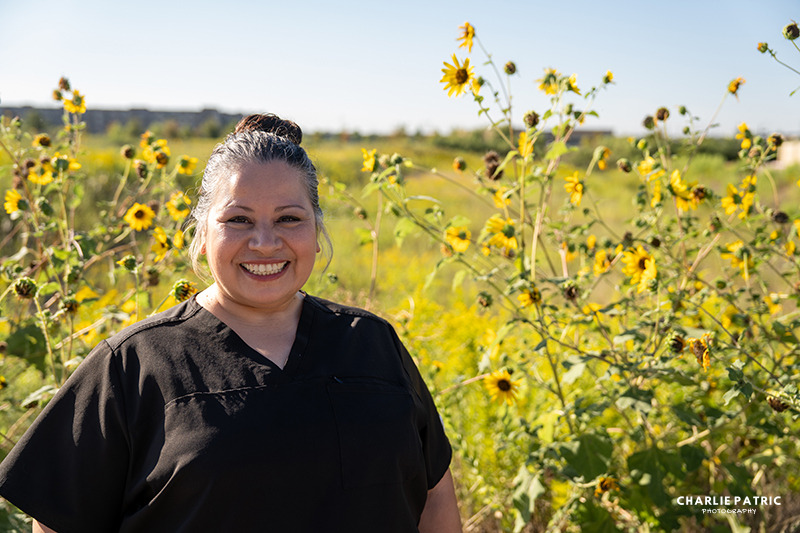 A woman in black scrubs smiles outdoors, standing in front of tall sunflowers and green vegetation under a clear sky—a great professional photo perfect for corporate headshots Frisco.