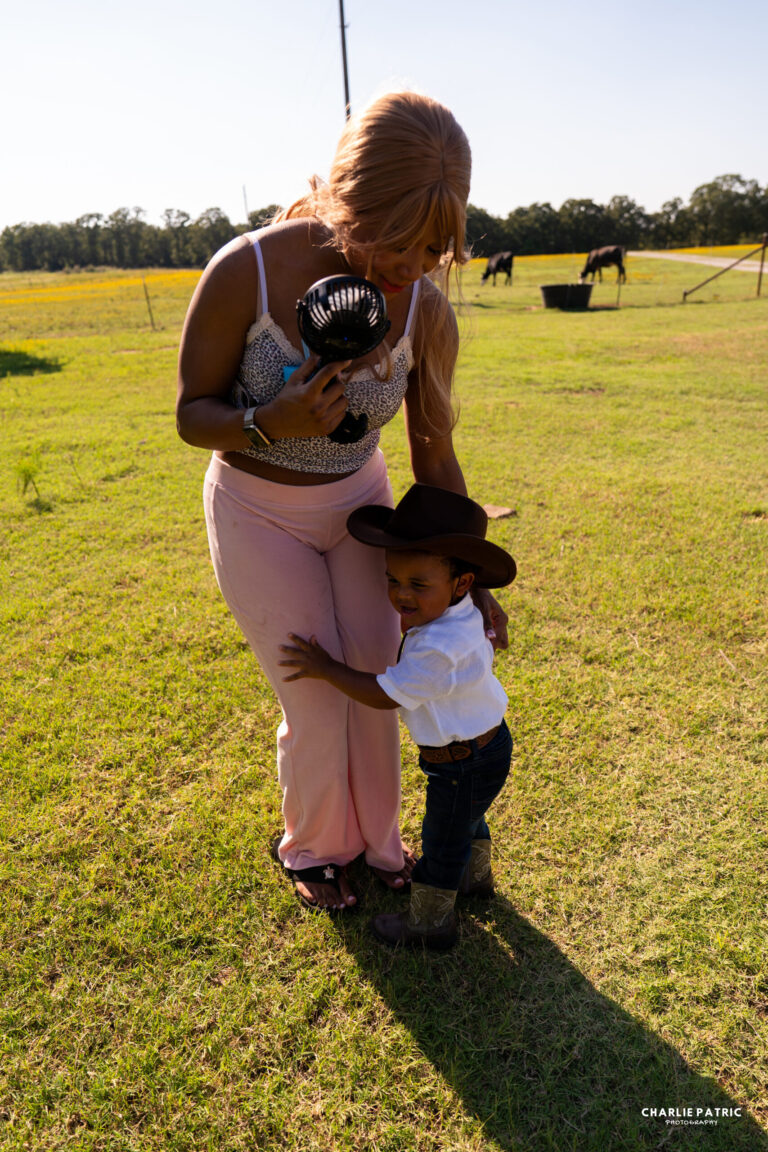 A woman stands on grass holding a fan while a young boy in a cowboy hat and boots hugs her leg; horses and trees create a picturesque backdrop, perfect for capturing sports or event photography tips in Frisco.