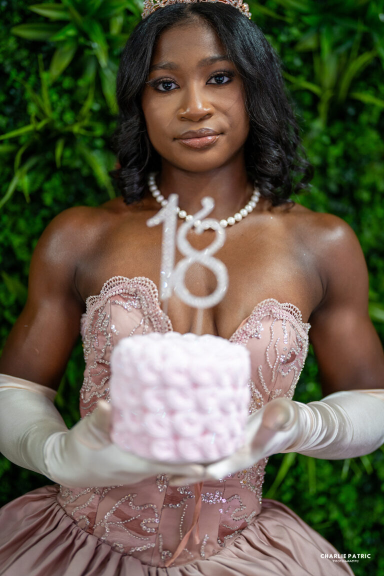 A young woman in a formal pink dress and gloves holds a small pink cake with an "18" topper, standing against a green leafy background—perfect inspiration for Frisco graduation photography services.