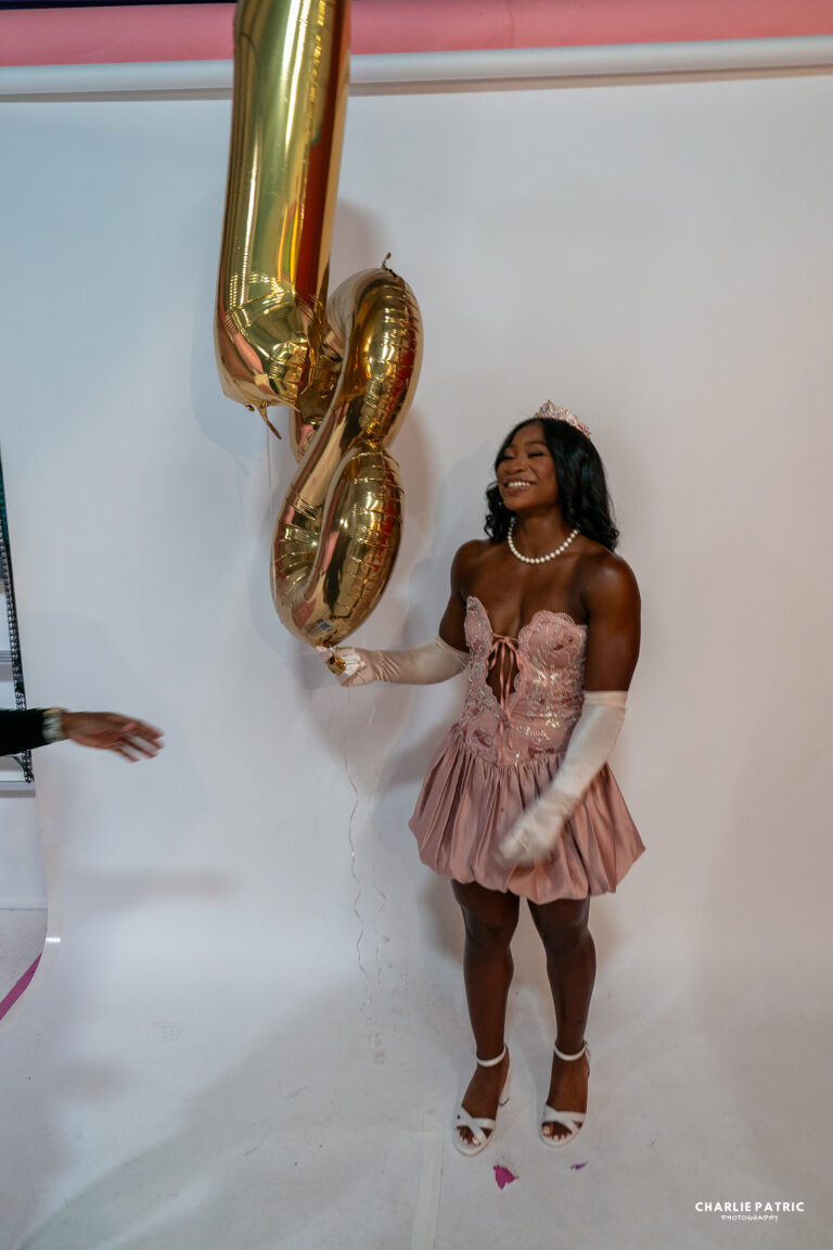 A young woman in a pink dress and tiara holds gold number balloons against a white backdrop, smiling—perfect for showcasing Frisco graduation photography services.