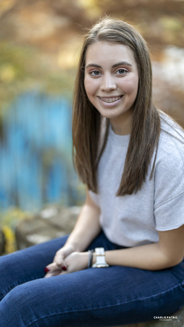 Young woman with straight brown hair, wearing a white t-shirt and jeans, sits outdoors on a stone bench, smiling at the camera—an ideal moment captured by Frisco graduation photography services. Background is blurred with hints of blue and greenery.