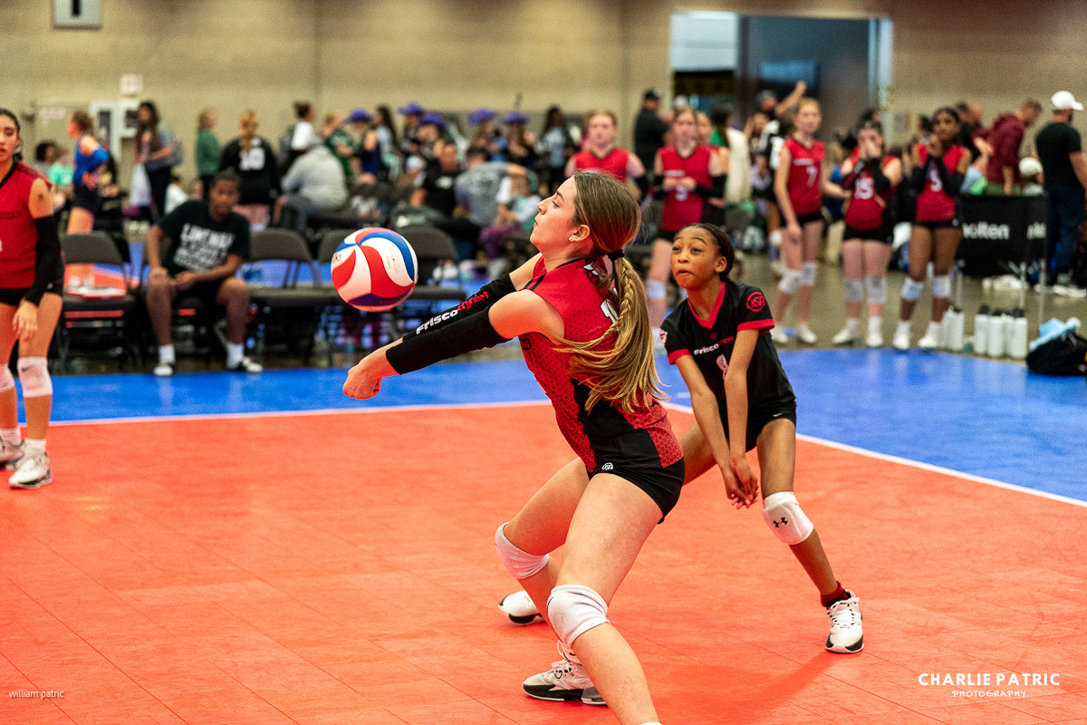 Two young volleyball players in red jerseys are ready to receive a serve in an indoor gym at a Frisco youth sports event, with teammates and spectators captured in the background, showcasing dynamic photography.