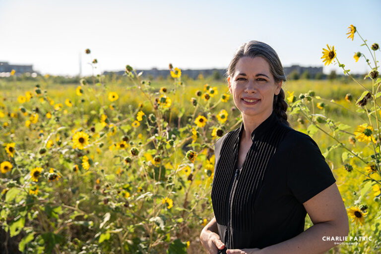 A woman with gray-streaked hair in a braid stands in a field of sunflowers, wearing a black top, as distant buildings—signs of urban life—appear under a clear sky. This could be the perfect setting for her new professional headshot.