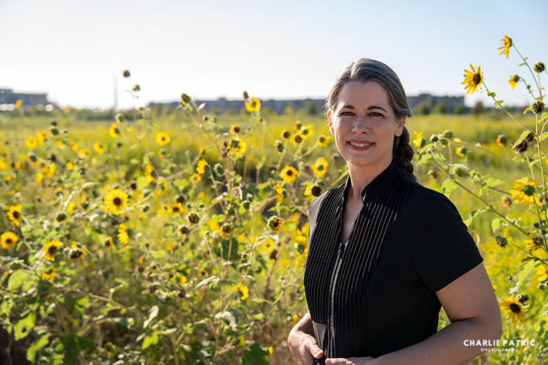 A woman with gray-streaked hair in a braid stands in a field of sunflowers, wearing a black top, as distant buildings—signs of urban life—appear under a clear sky. This could be the perfect setting for her new professional headshot.